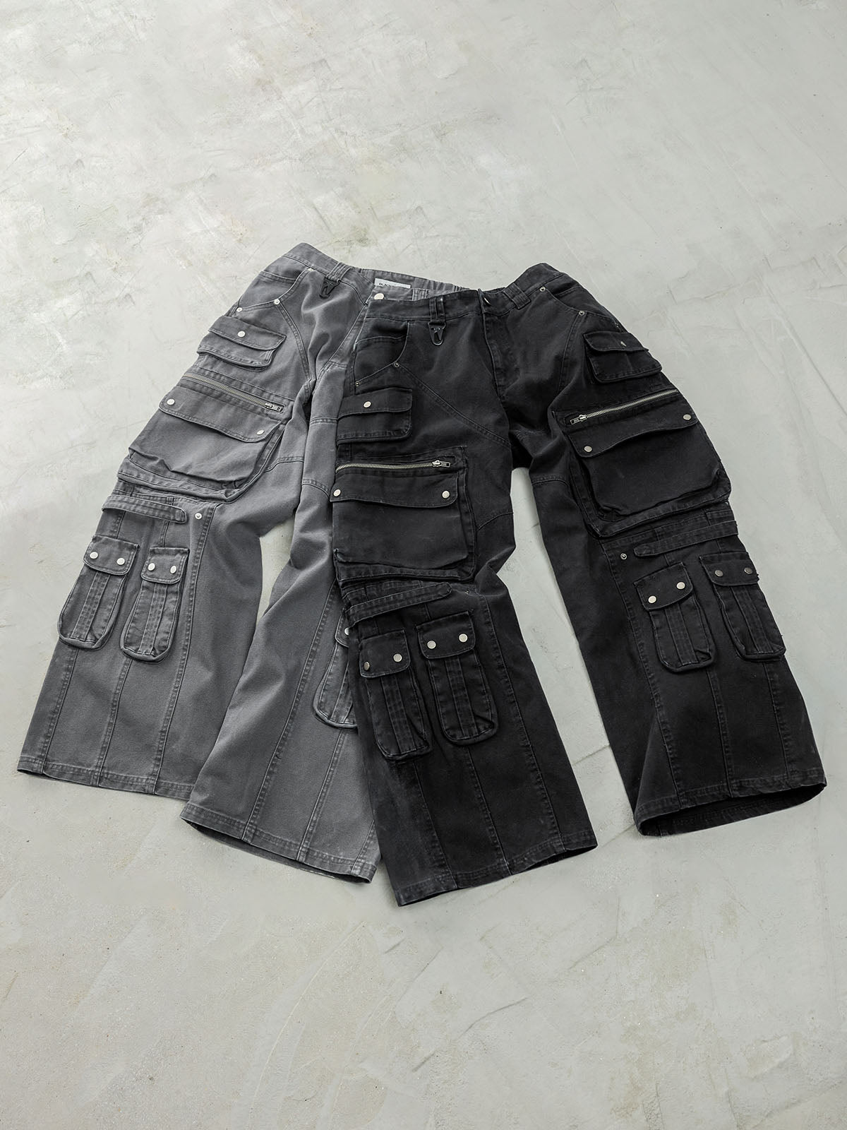 Cargo pants laid out on a concrete surface, one pair in grey and the other in black, both featuring multiple large pockets, zippers, and snap-button details.