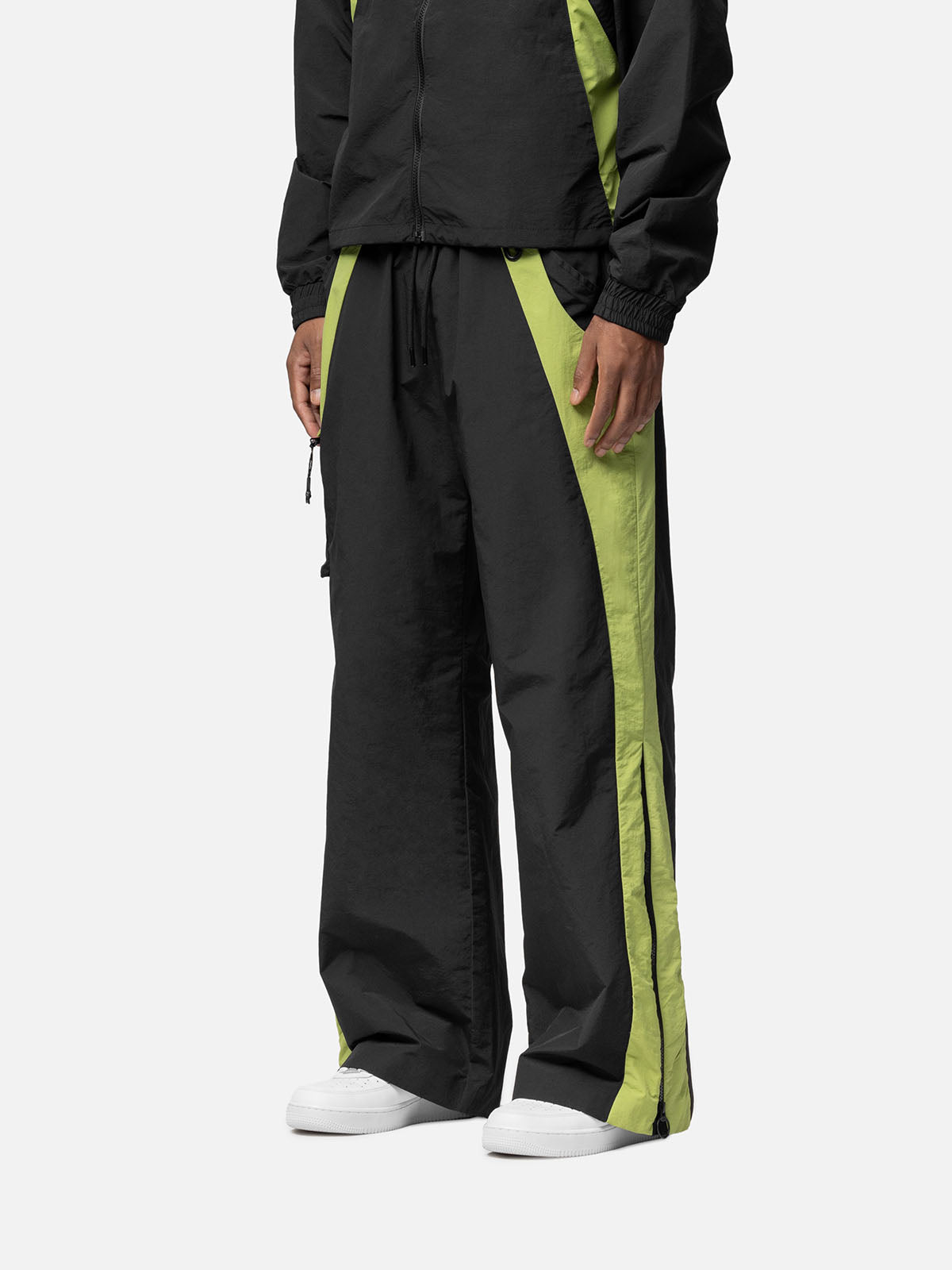 Side view of a black with green side panels track pants , decorative buckle at the waist, matching jacket, and white sneakers.