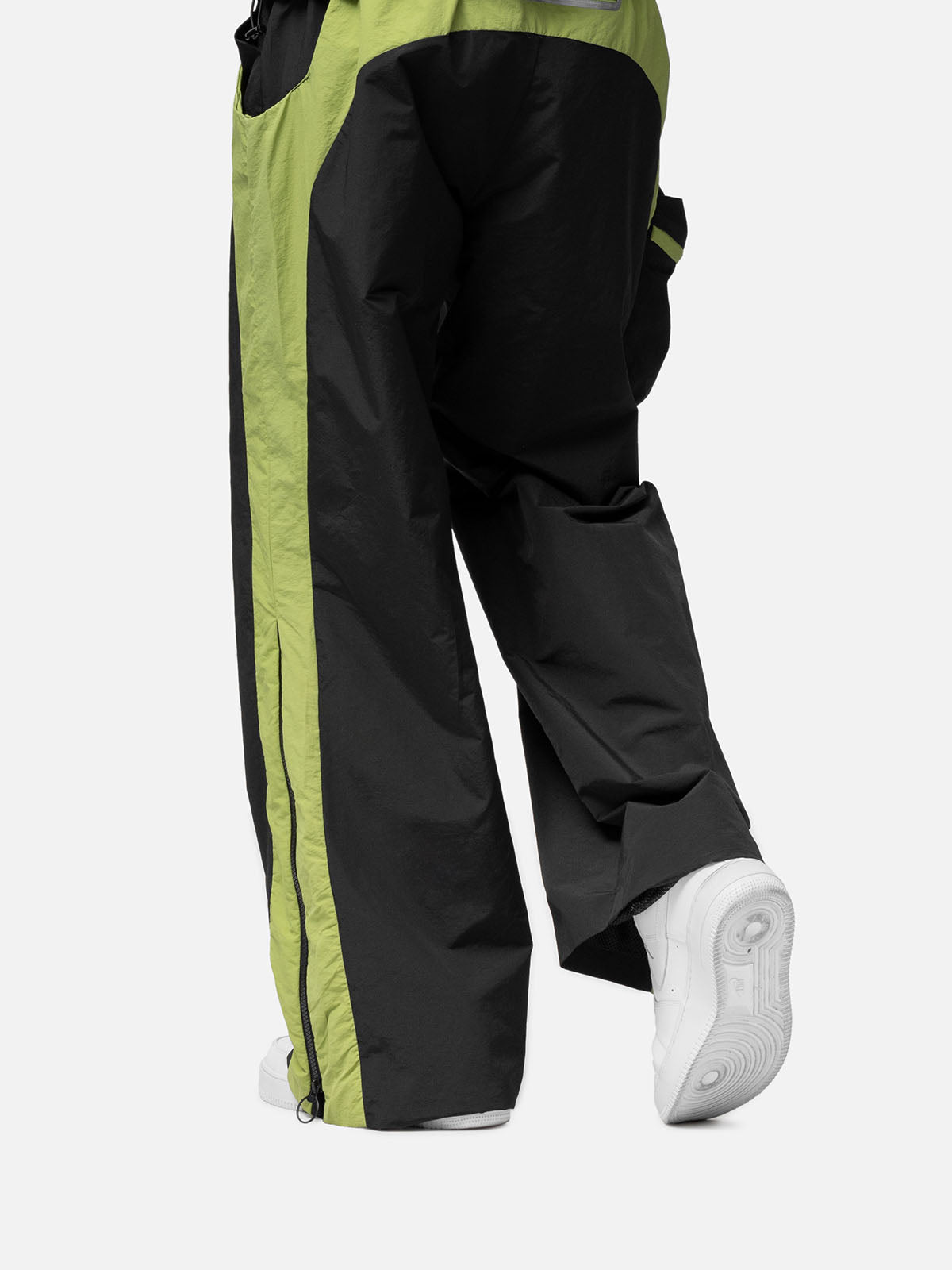 Close-up of a black with green side panels track pants , a zip-up expandable seam to hem, paired with white sneakers.