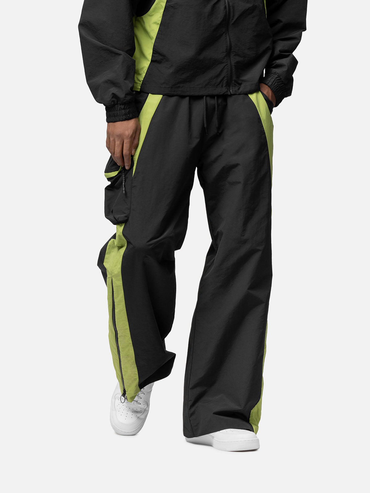 Front view of a black with green side panels track pants, with side pocket on the right side, decorative buckle at the waist, matching jacket, and white sneakers.