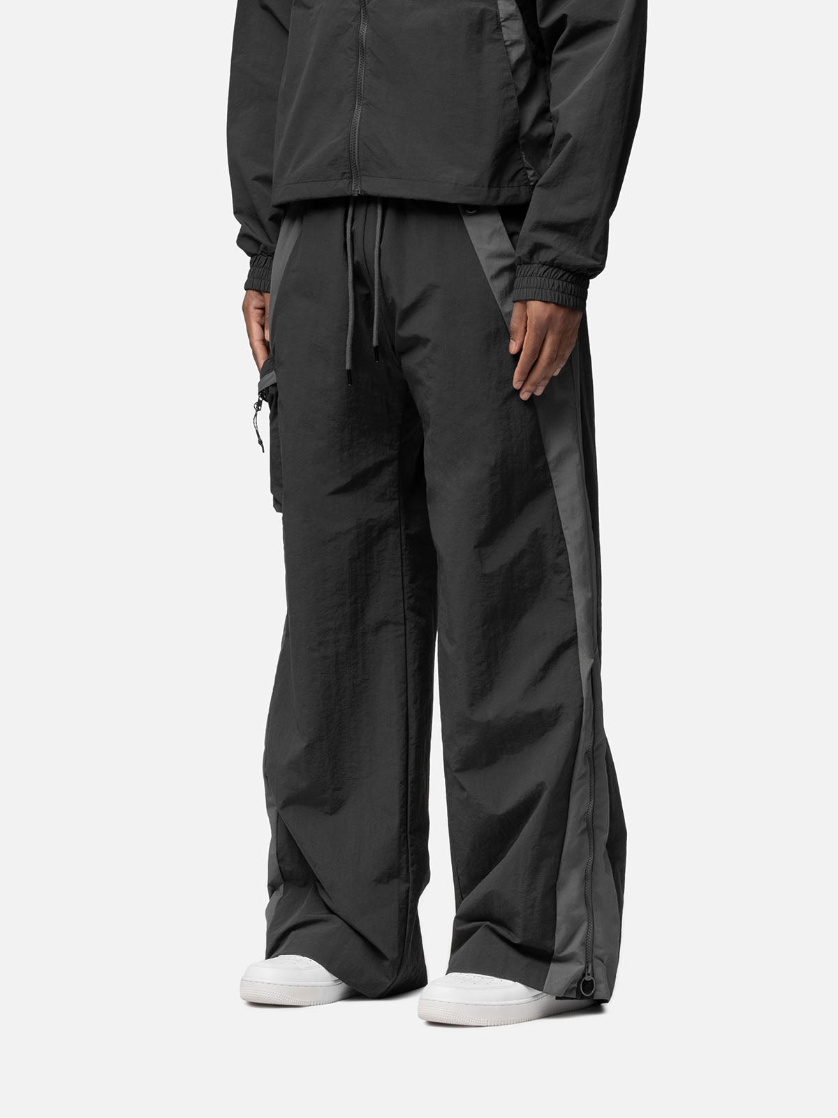 Side view of a charcoal with grey side panels track pants , decorative buckle at the waist, matching jacket, and white sneakers.
