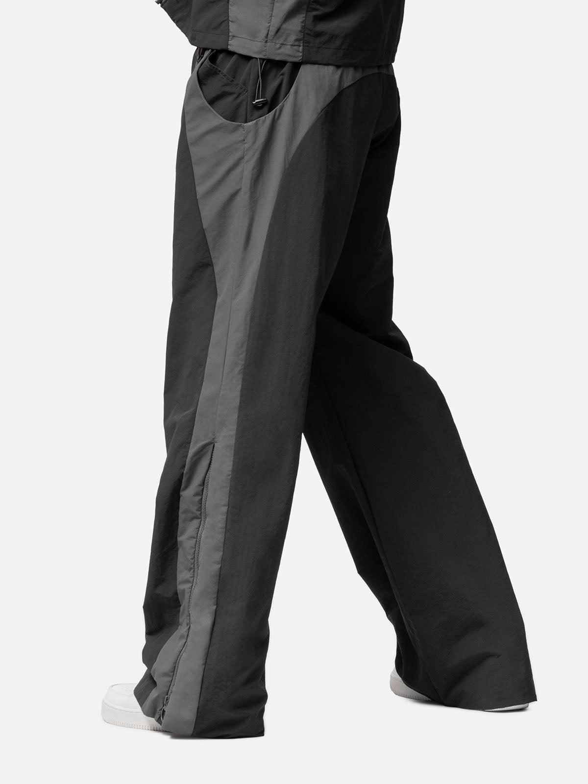 Side view of a charcoal with grey side panels track pants , decorative buckle at the waist, matching jacket, and white sneakers.