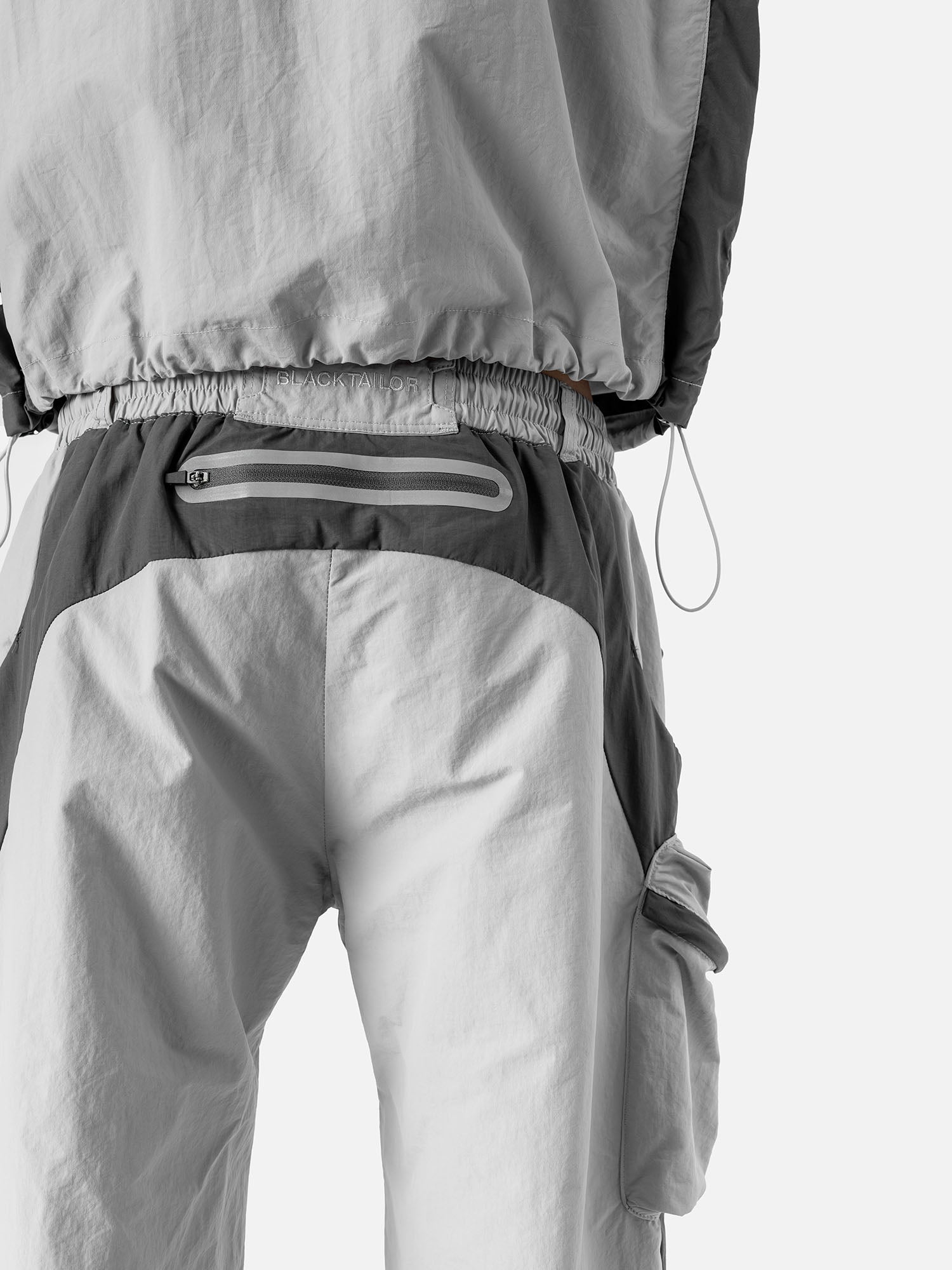 Close-up of the back pocket of a light grey with grey side panels track pants, featuring a brand logo embroidery at the waist.