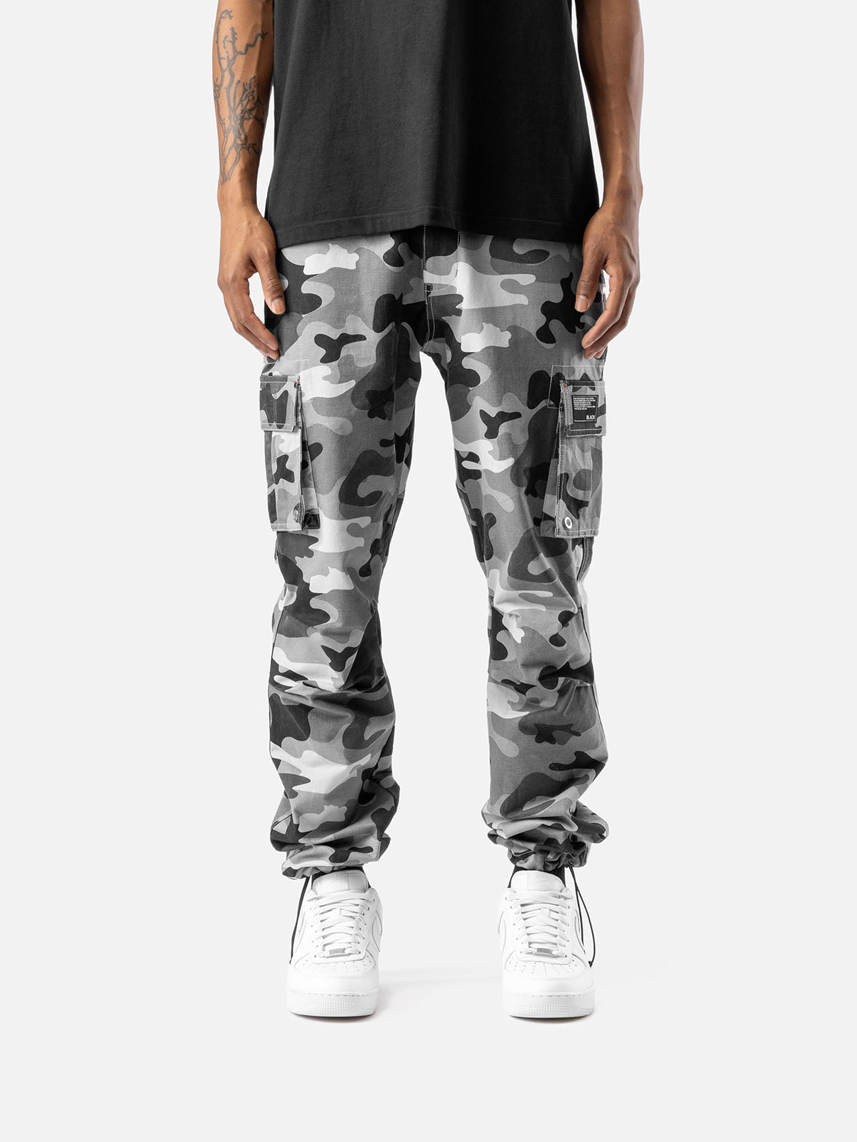 Grey camo cargo pants with side pockets, ankle drawstrings, paired with a black t-shirt, and white sneakers, showcasing streetwear style.