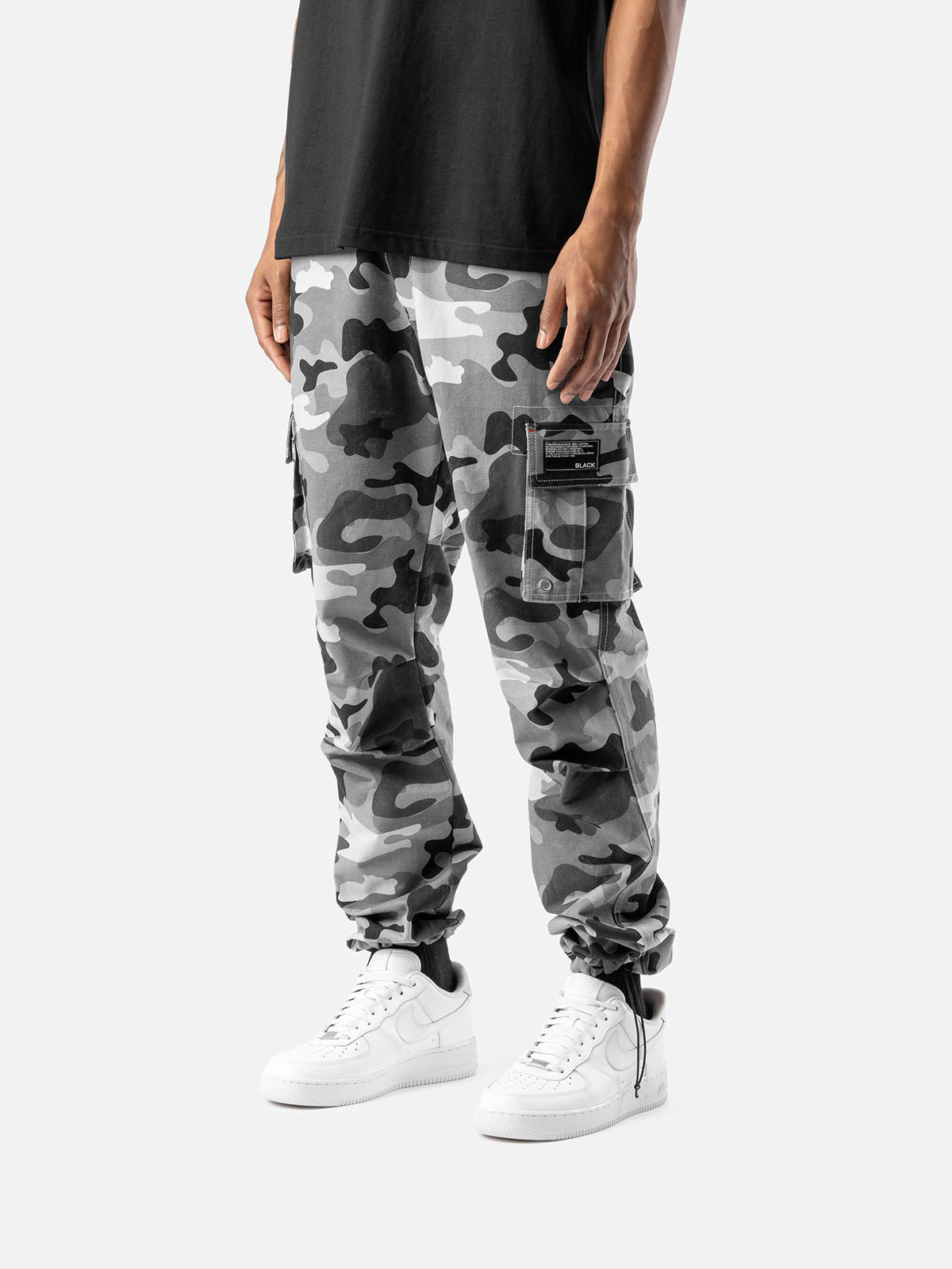 Side view of grey camo cargo pants with side pockets, ankle drawstrings, paired with a black t-shirt, and white sneakers.