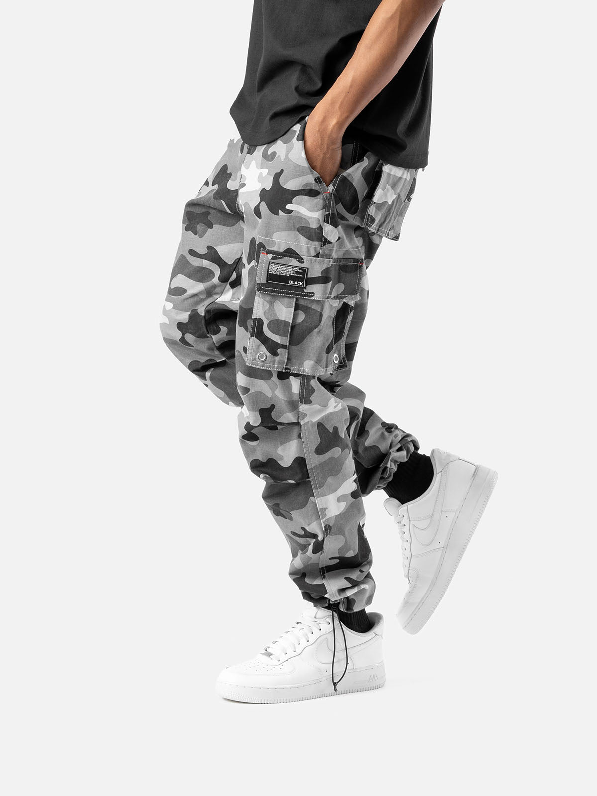 Side view of grey camo cargo pants with side pockets, ankle drawstrings, paired with a black t-shirt, and white sneakers.