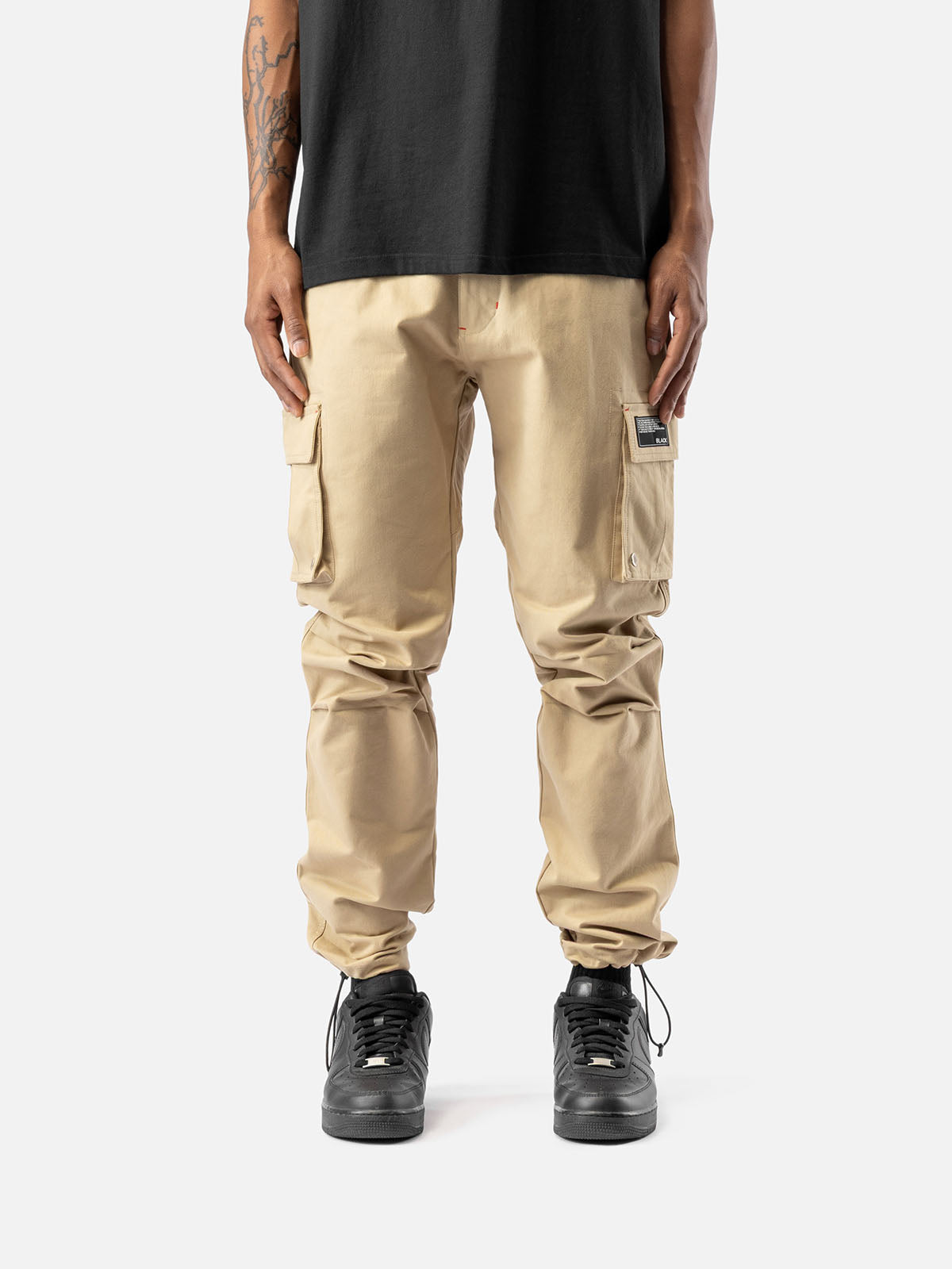 Khaki cargo pants with side pockets, ankle drawstrings, paired with a black t-shirt, and black sneakers, showcasing streetwear style.