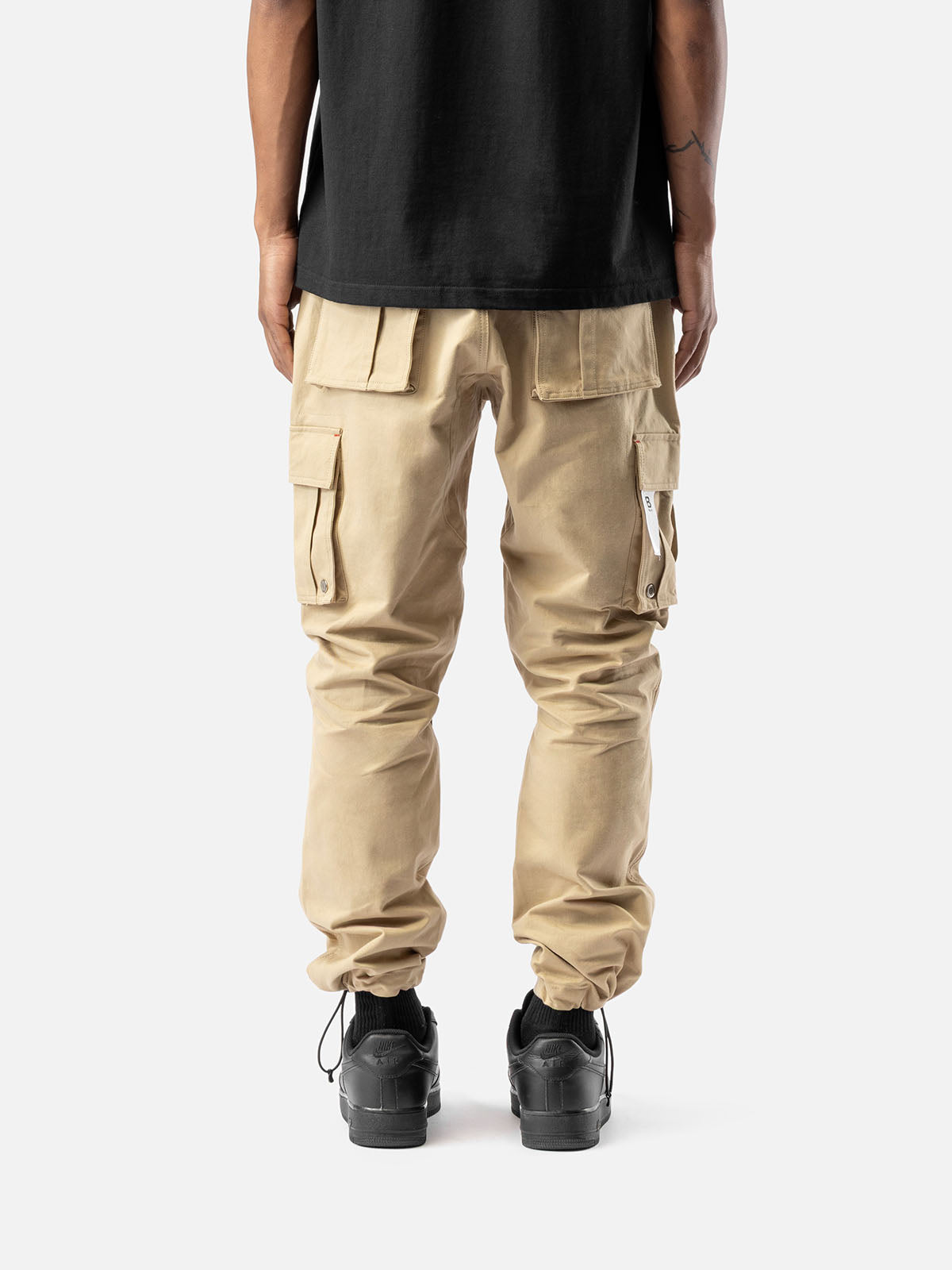 Back view of khaki cargo pants with side pockets, ankle drawstrings, and simple back pockets, paired with a black t-shirt, and black sneakers.