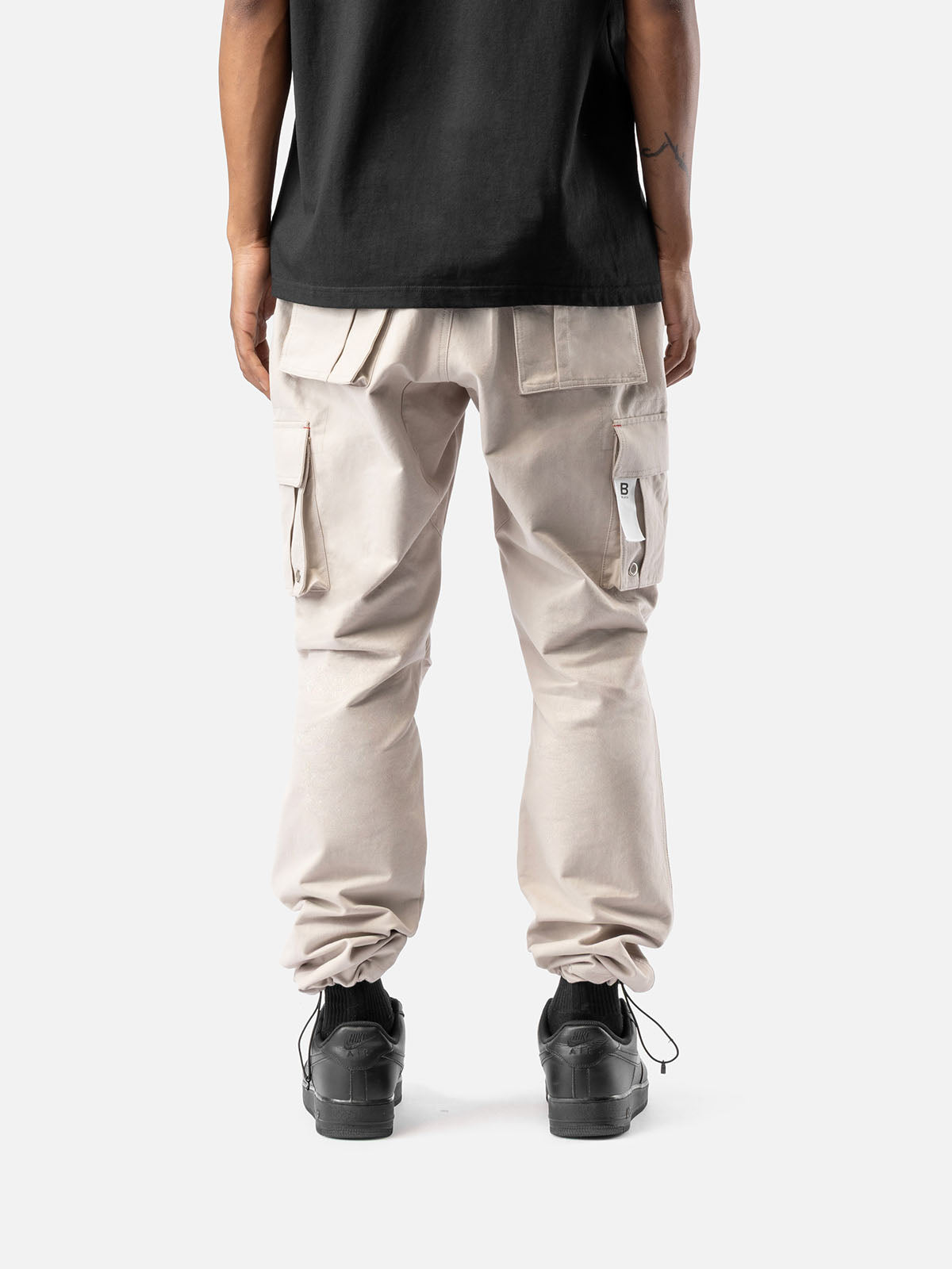 Back view of rock cargo pants with side pockets, ankle drawstrings, and simple back pockets, paired with a black t-shirt, and black sneakers.