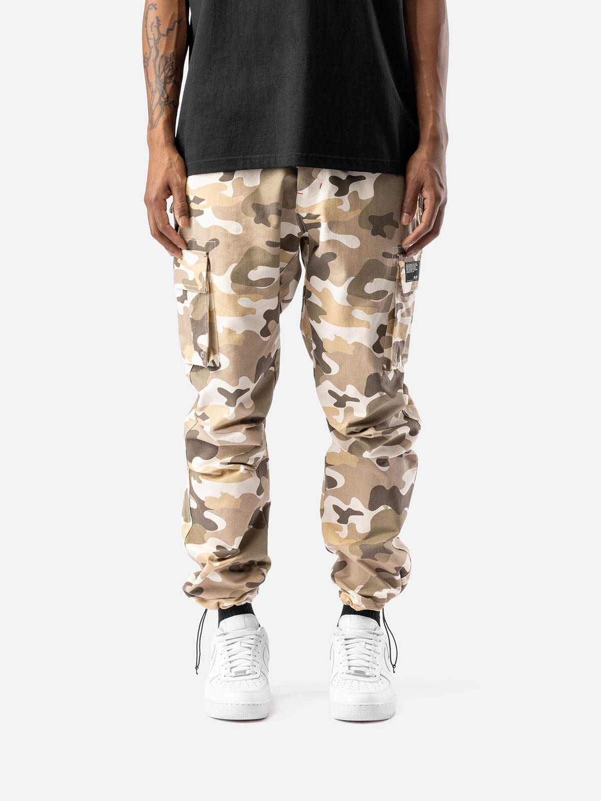 Sand camo cargo pants with side pockets, ankle drawstrings, paired with a black t-shirt, and white sneakers, showcasing streetwear style.