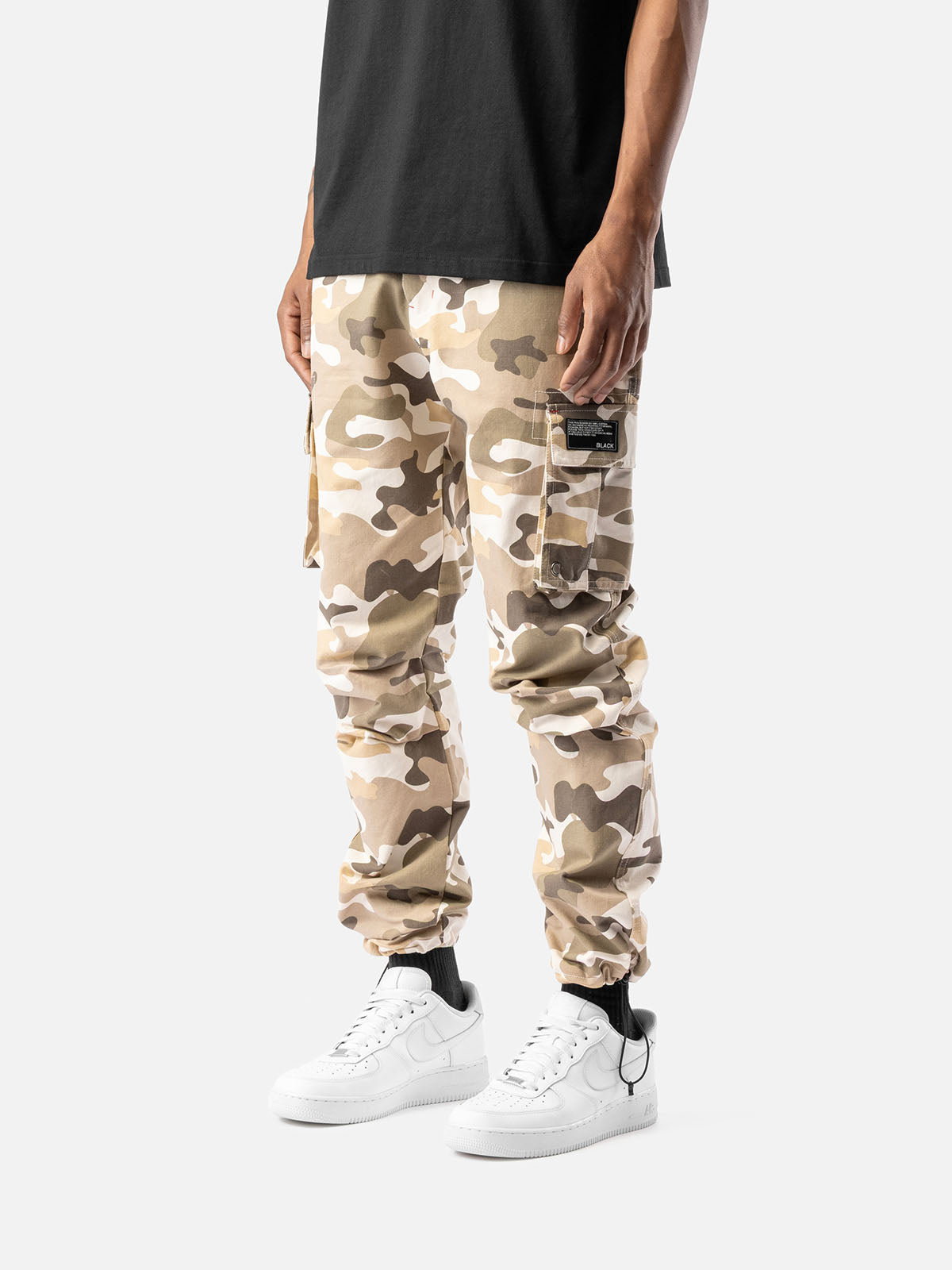Side view of sand camo cargo pants with side pockets, ankle drawstrings, paired with a black t-shirt, and white sneakers.
