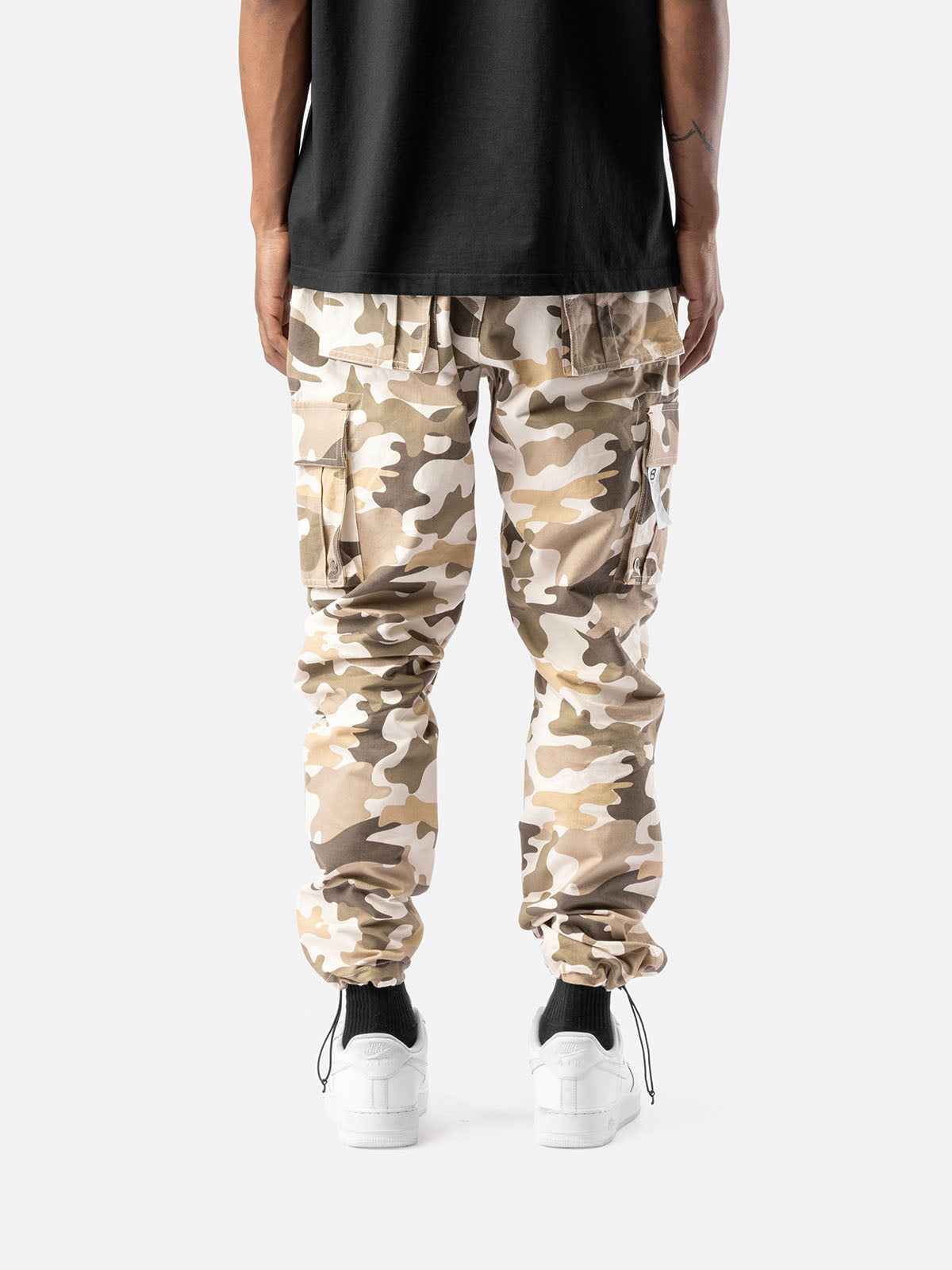 Back view of sand camo cargo pants with side pockets, ankle drawstrings, and simple back pockets, paired with a black t-shirt, and white sneakers.