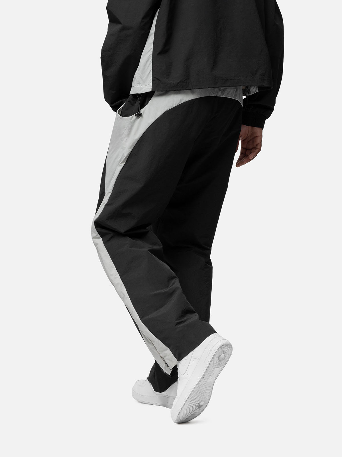 Back view of black with white side panels track pants, a zip-up expandable seam to hem, matching jacket, and white sneakers.