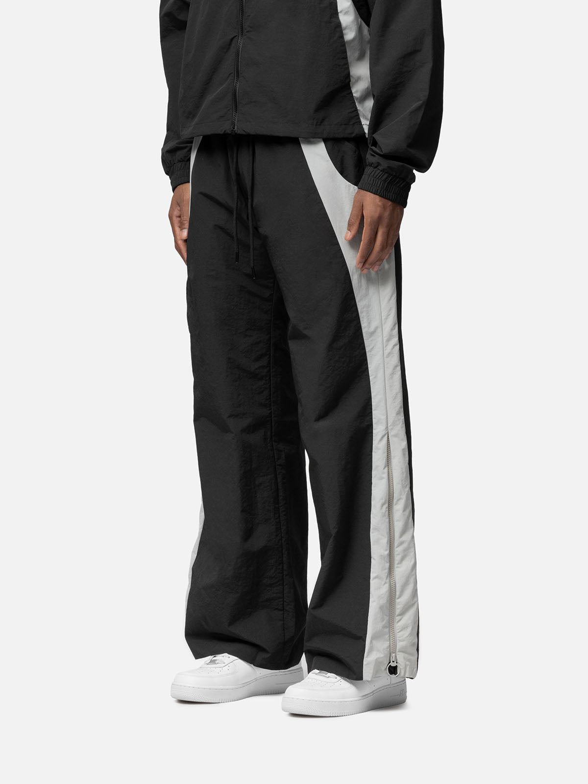 Side view of a black with white side panels track pants , decorative buckle at the waist, matching jacket, and white sneakers.